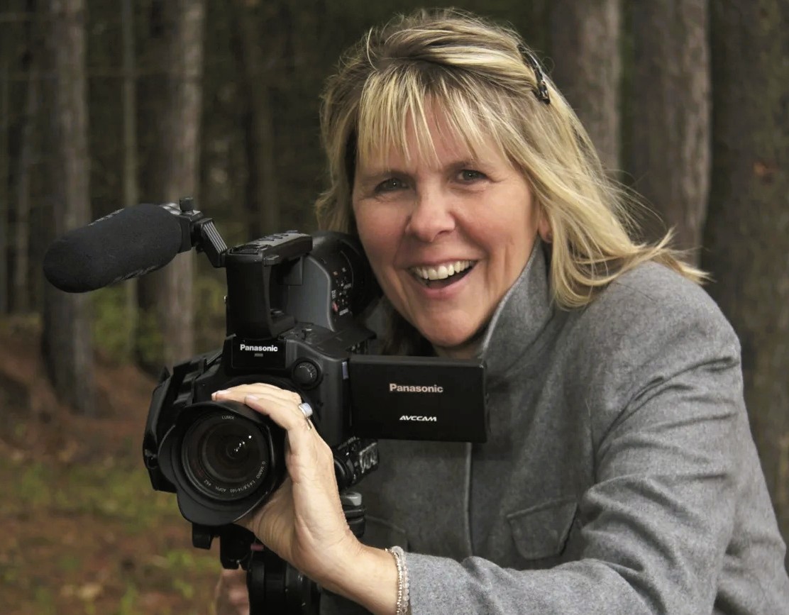A woman outdoors holds a Panasonic video camera, smiling at the camera, with trees visible in the background.