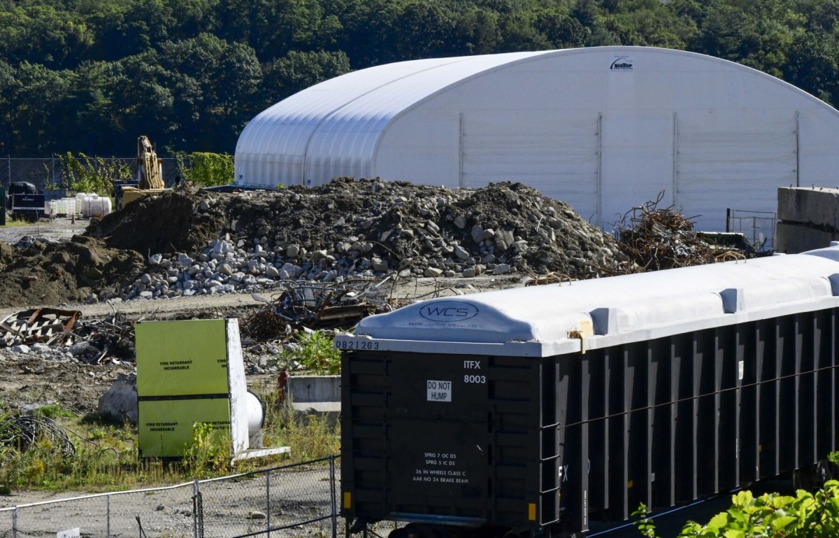 A freight train car sits on tracks in front of a pile of rubble and debris, with a white industrial tent and construction equipment in the background.