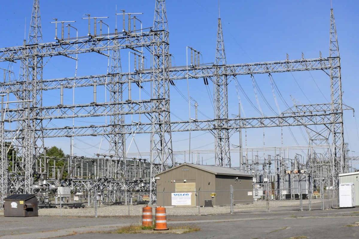 Electrical substation with metal towers, wires, a small building, and orange traffic barrels in front, surrounded by a chain-link fence under a clear sky.