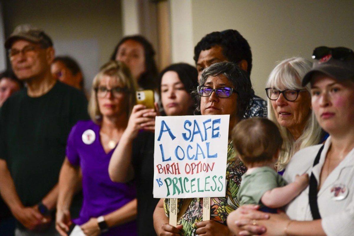 A group of people stands indoors; one person in the center holds a sign reading, “A safe local birth option is priceless.”.
