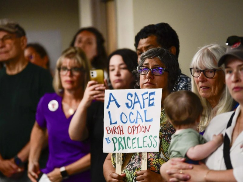 A group of people stands indoors; one person in the center holds a sign reading, “A safe local birth option is priceless.”.