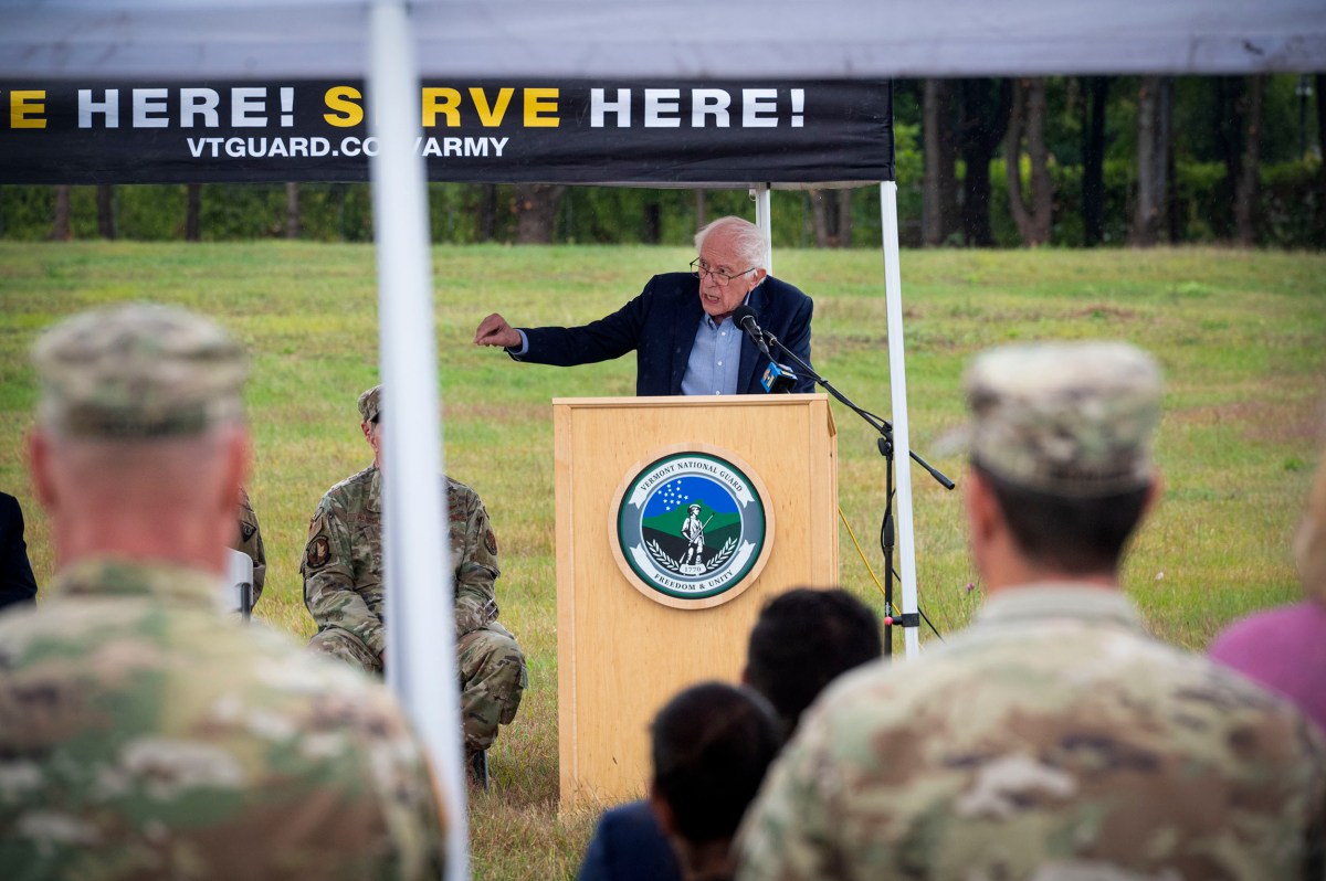 A man speaks at a podium outdoors under a tent, with military personnel seated and standing nearby. The podium has an insignia and a banner reads, “SERVE HERE! VTGUARD.COM/ARMY.”.