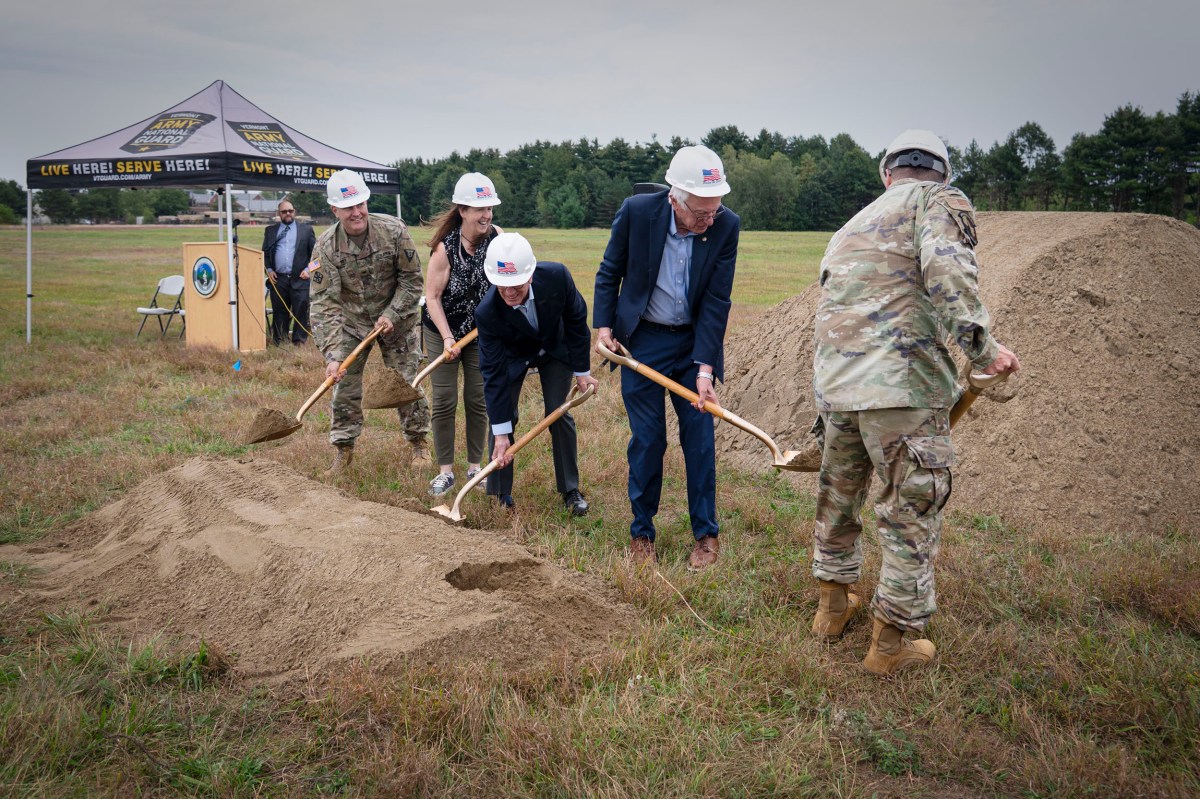 Five people in hard hats, including military personnel and civilians, participate in a groundbreaking ceremony, shoveling dirt at an outdoor site with a tent and piles of soil nearby.