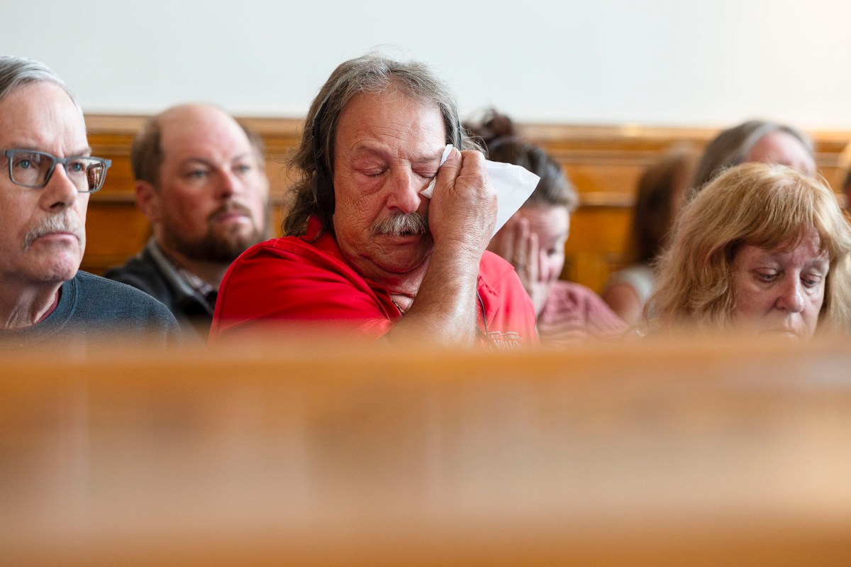 A man in a red shirt wipes his eye with a tissue while sitting among others in a courtroom.