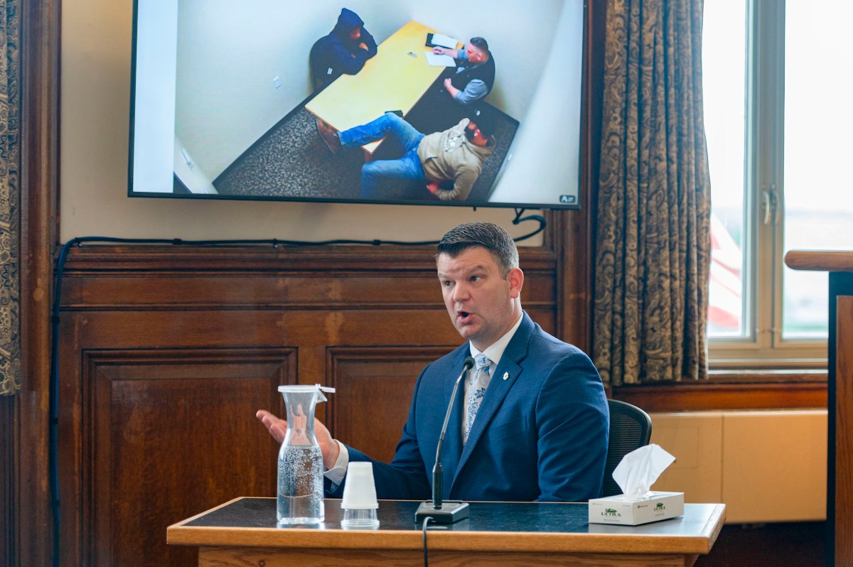 A man in a suit testifies in court; behind him, a screen shows a video of two people in an interview room.