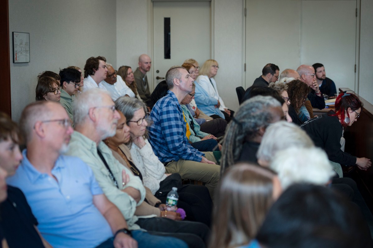 A group of people sit closely together in a conference room, listening attentively to a speaker who is out of frame.