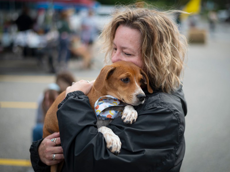 A woman with wavy blonde hair hugs a brown and white puppy wearing a patterned bandana at an outdoor event.
