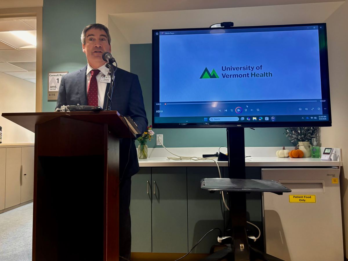 A man in a suit speaks at a podium next to a screen displaying the University of Vermont Health logo in a room with cabinets and a small pumpkin decoration.