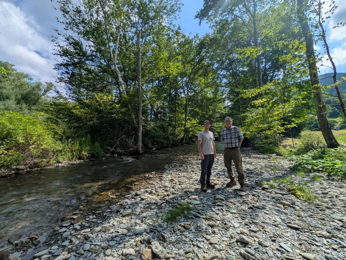 Two people stand on a rocky riverbank surrounded by trees and greenery under a blue sky with clouds.