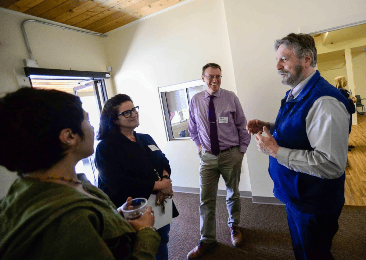 Four adults stand indoors in conversation; one person gestures while the others listen, all dressed in business-casual attire.