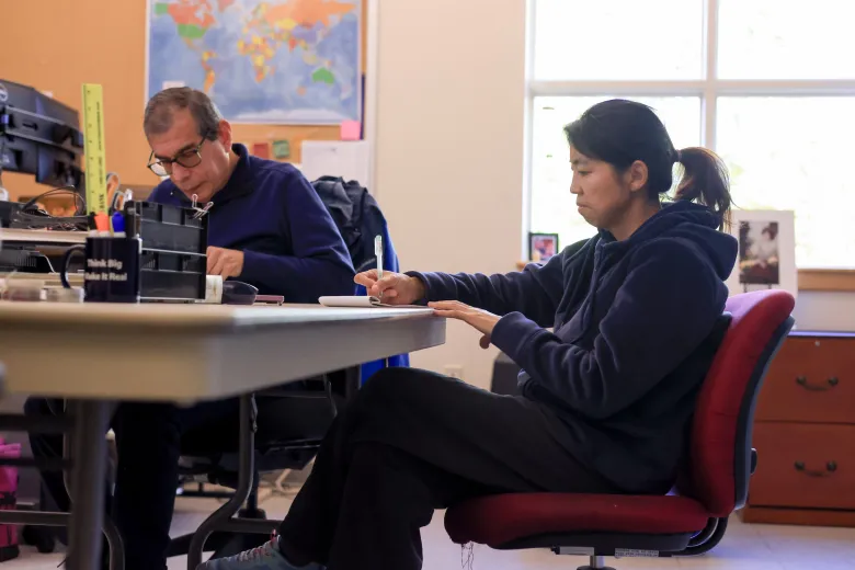 Two people sit at a desk in an office, one writing in a notebook and the other working on a computer.