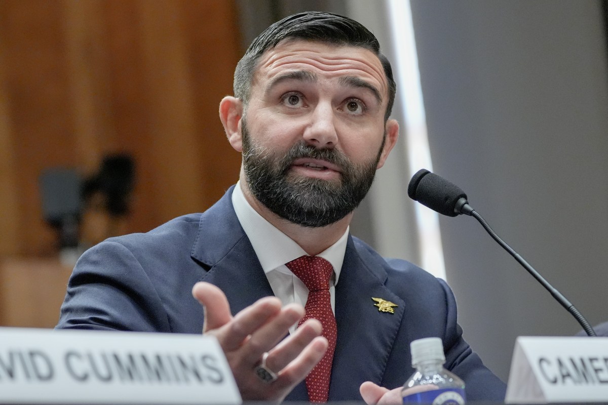 A bearded man in a suit and tie speaks into a microphone at a table with nameplates.
