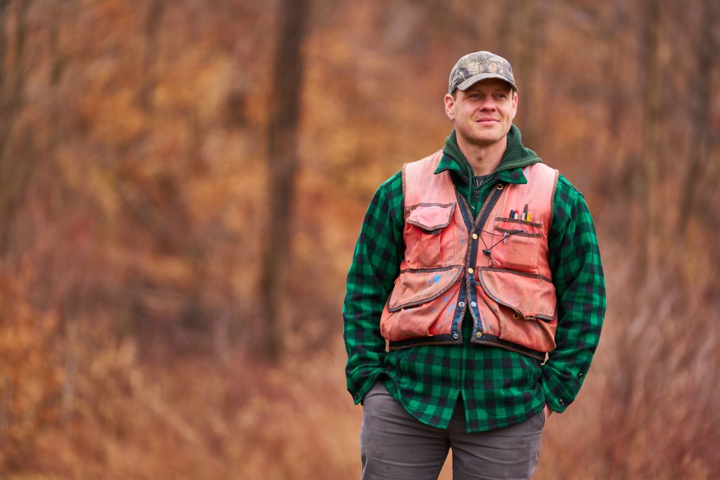 A man in a green flannel shirt and orange hunting vest stands outdoors.