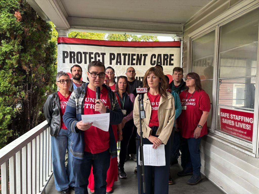 A group of nurses in “Union Strong” t-shirts stands on a porch behind two speakers holding microphones and papers in front of a “Protect Patient Care” banner.