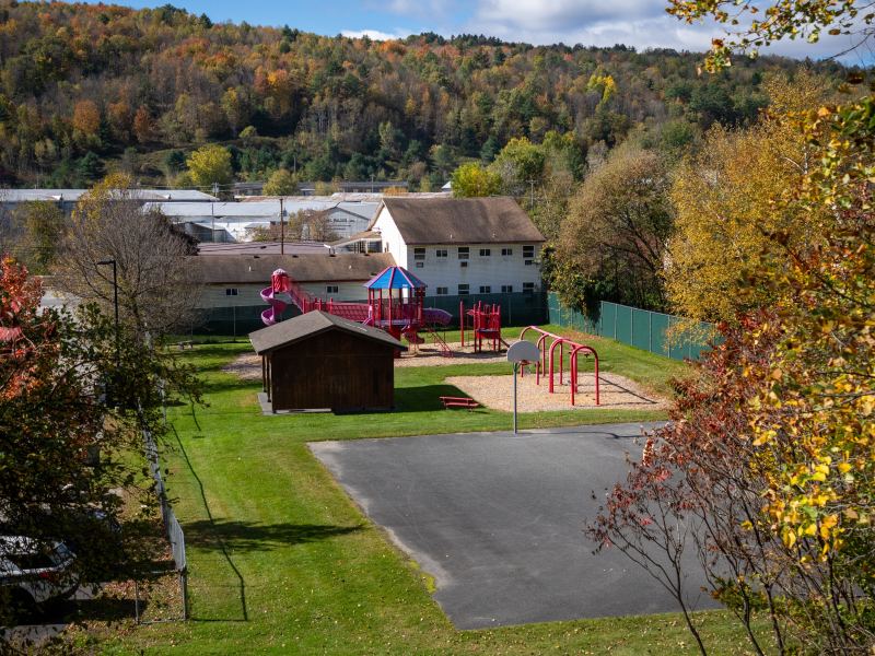 A playground with a slide, climbing structure, and swings next to an asphalt court, set against a backdrop of autumn trees and buildings.