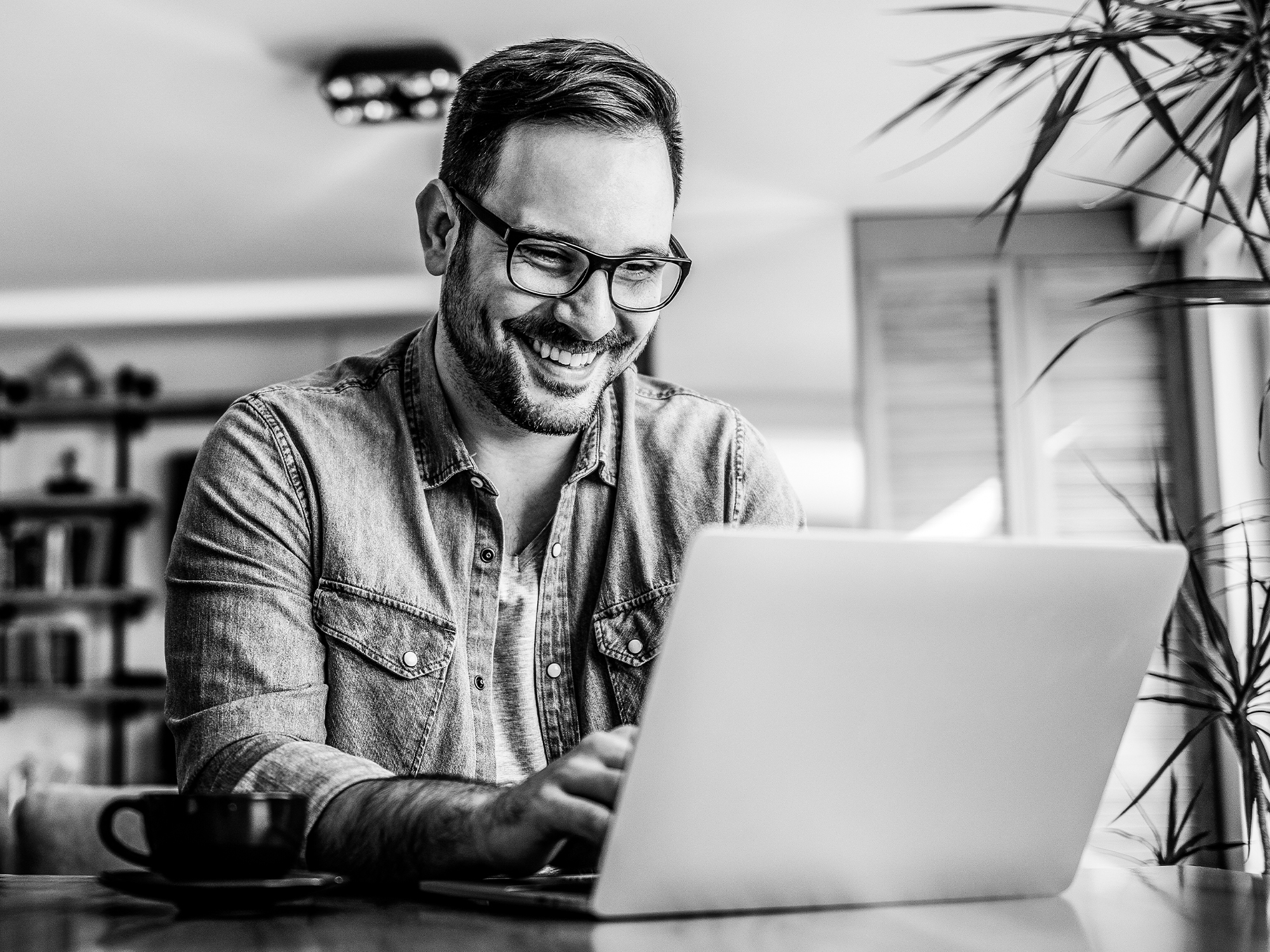 A smiling man with glasses sits at a table using a laptop.