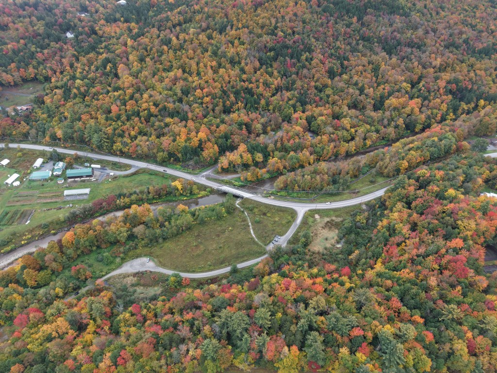 Aerial view of a road intersecting a forested area with autumn foliage and a small bridge over a stream.