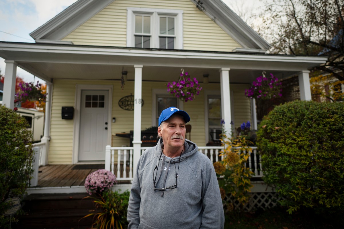 A man in a gray hoodie and blue baseball cap standing in front of a yellow house with a covered porch.