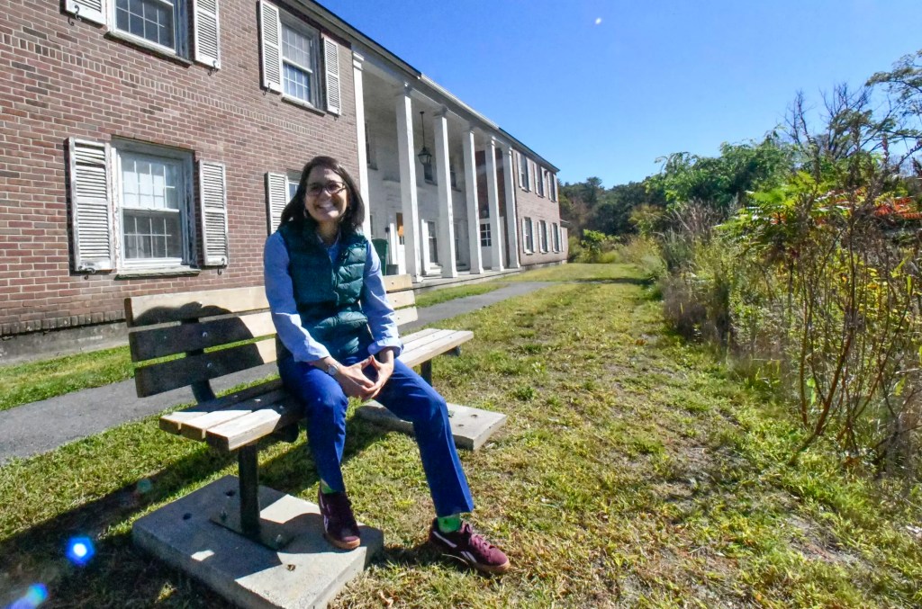 A smiling woman in a vest and glasses sits on a wooden park bench in front of a brick building with white columns.