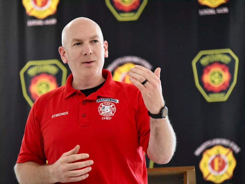 Brattleboro Fire Chief Symonds in a red polo shirt speaking in front of a backdrop with fire department logos.