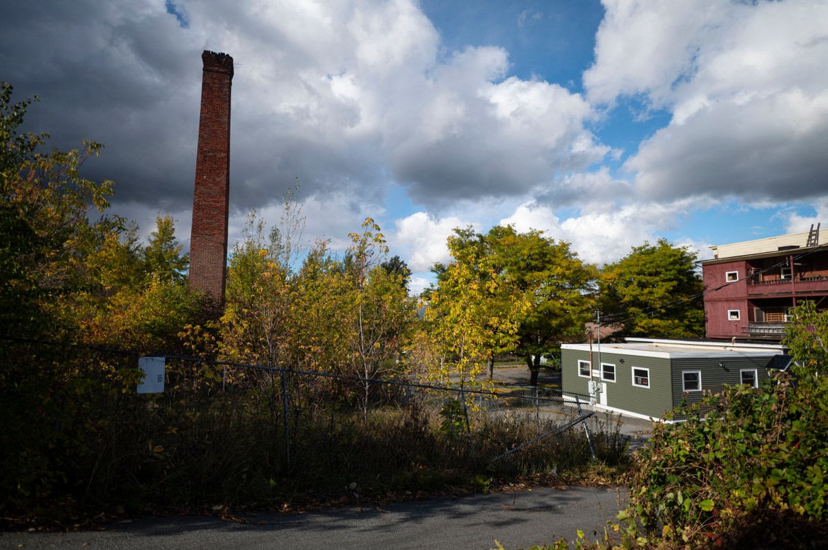 A tall brick smokestack rises above trees and a small portable office building behind a chain-link fence.