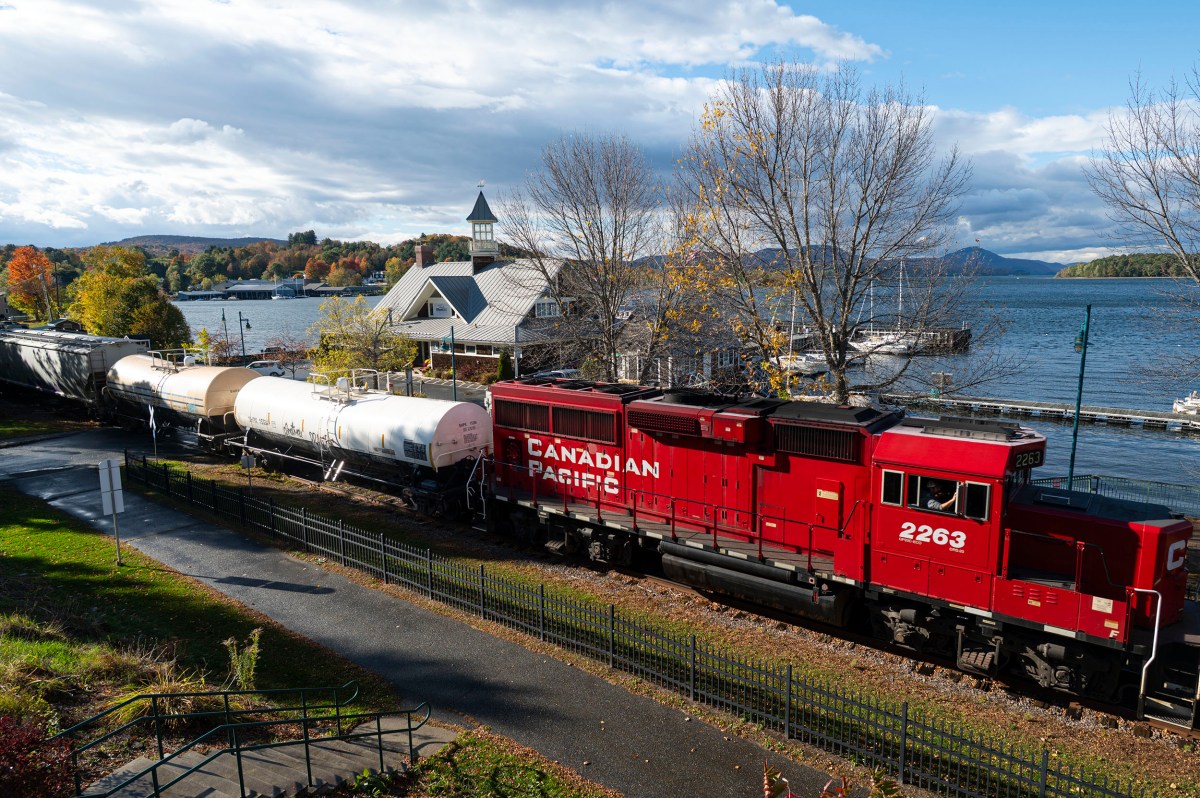 A red Canadian Pacific freight train travels along tracks beside a lake, with a building in the background.