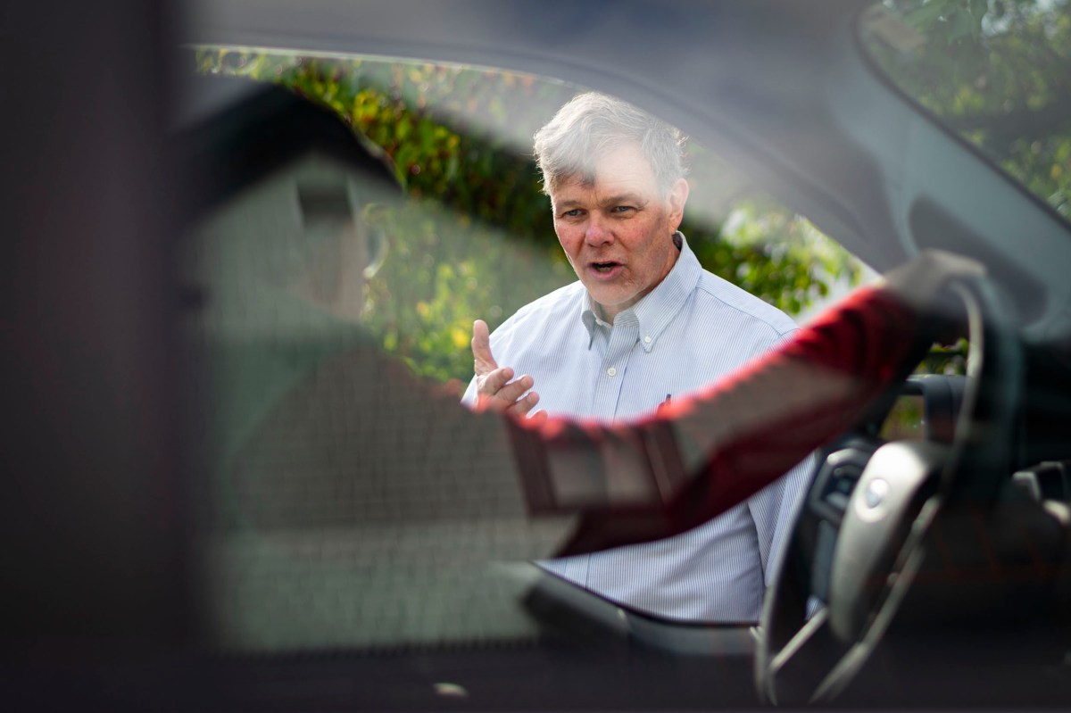 A man in a button-down shirt gesturing while seen through a car window.