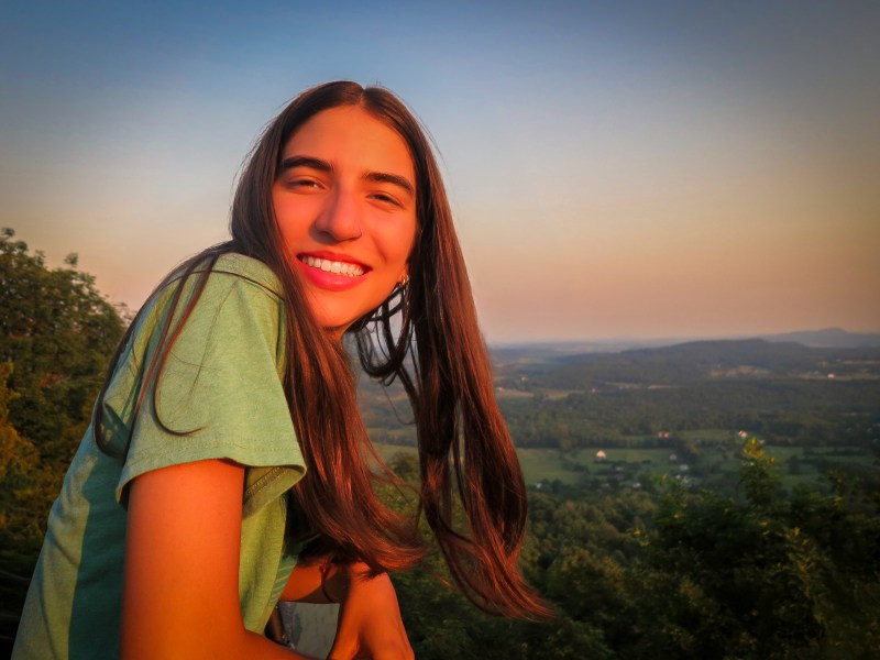 A young person smiling at the camera with a vast, hilly landscape in the background during sunset.